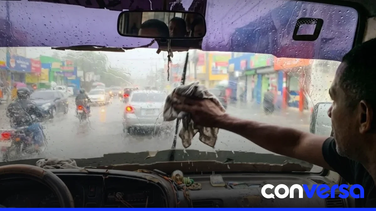 Imagen de una guagua pública con puertas abiertas y sin limpiavidrios durante una lluvia.